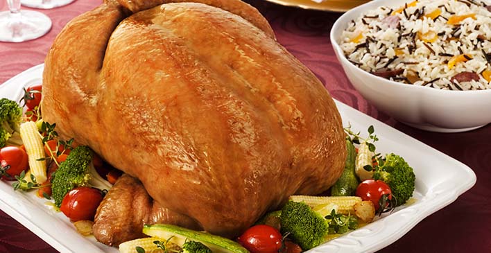 Prato retangular branco com milho, pepino, brócolis, tomate, tomilho e um frango inteiro assado, ao lado uma tigela com arroz