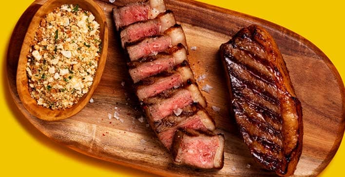 tábua de madeira oval com uma tigela de madeira em cima preenchida com farofa e dois pedaços de bife, um deles está fatiado