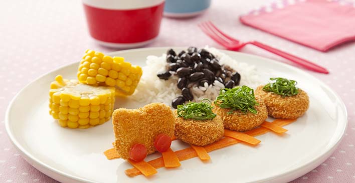 Prato branco com dois pedaços de uma espiga de milho, arroz com feijão preto em cima, quatro nuggets simulando um trem com cenoura simulando os trilhos de um trem, tomate simulando as rodas do trem e couve