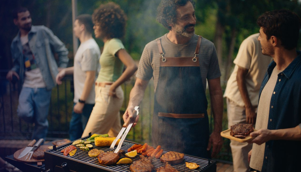 Homem grelhando e sorrindo para amigos em churrasco.