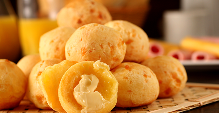 diversos pães de queijo e um deles aberto ao meio com manteiga, ao fundo alguns enrolados de presunto e queijo e um jarro de suco de laranja
