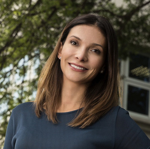 Marcela Rocha com uma blusa azul sorrindo para a câmera com uma arvore ao fundo