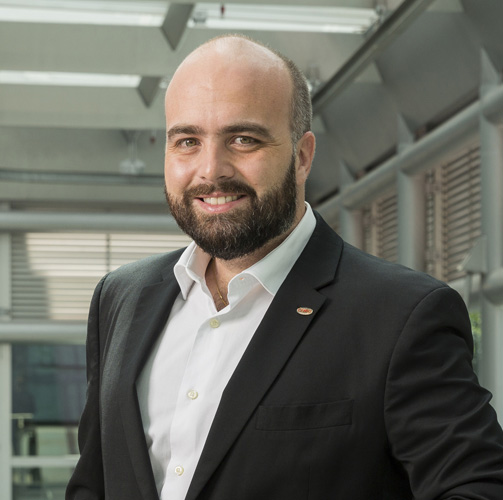 Guilherme Motta com um terno preto e uma camisa social branca sorrindo para a câmera com um escritório ao fundo