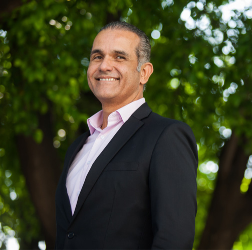 Renato Costa com um terno preto e uma camisa social rosa sorrindo para a câmera com uma arvore ao fundo