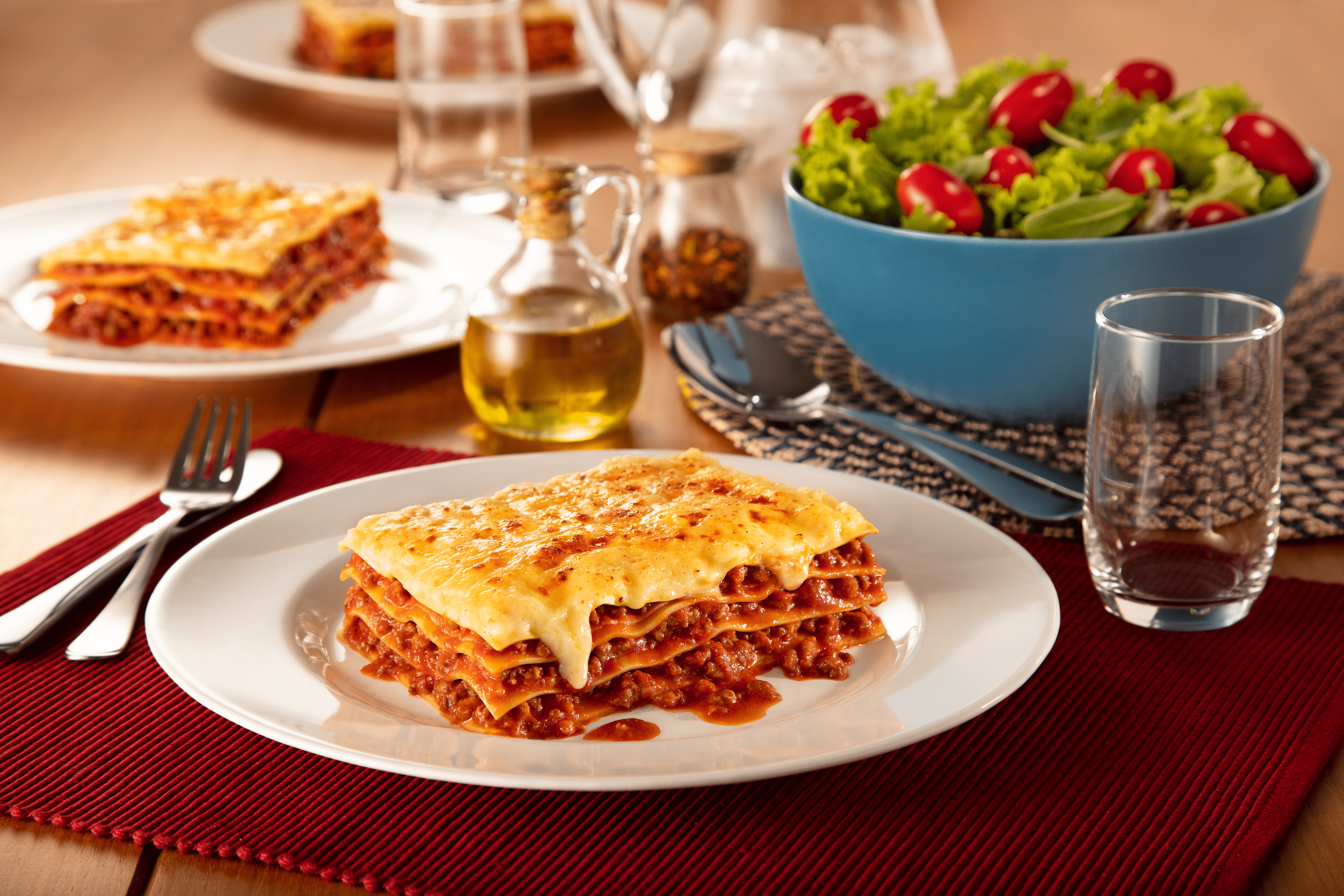 mesa posta com dois pratos brancos de lasanha, dois frascos um com tempero e um com azeite e uma tigela de salada
