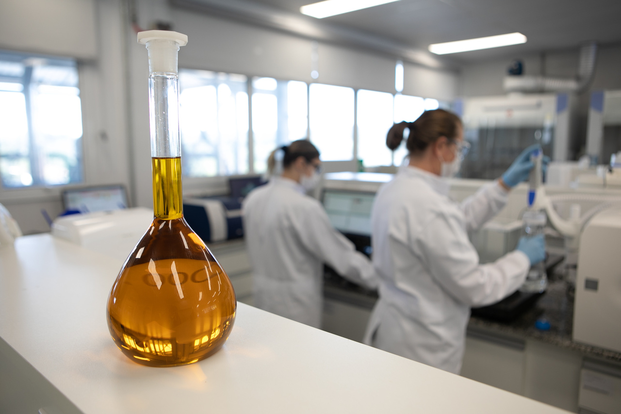 laboratório com um balão volumétrico e duas mulheres ao fundo vestindo jalecos, óculos de proteção e mascaras