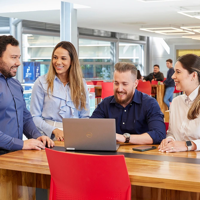 Four coworkers talk around a laptop in an office setting.