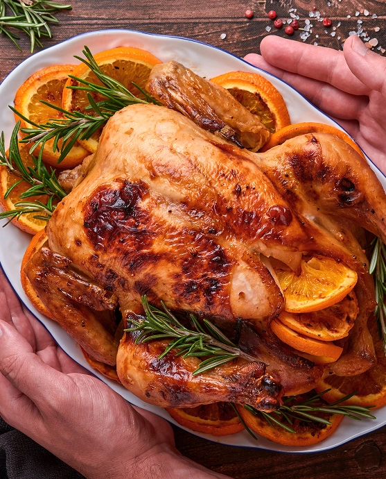 Whole roasted chicken on a platter, garnished with orange slices and rosemary sprigs on a wooden table.
