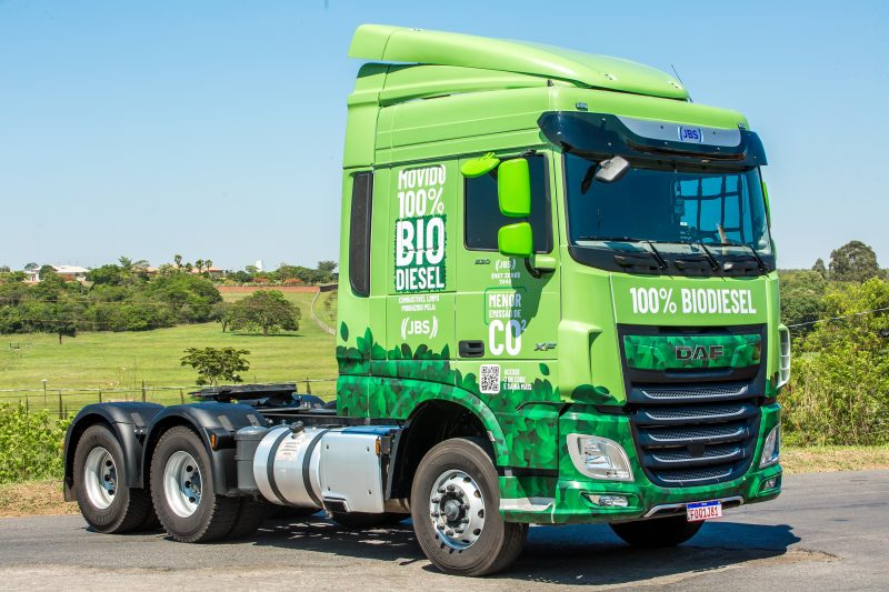 Caminhão de carga verde estacionado em uma estrada, com adesivos destacando o uso de 100% biodiesel. Ao fundo, há uma paisagem rural com gramado, árvores e céu azul aberto.