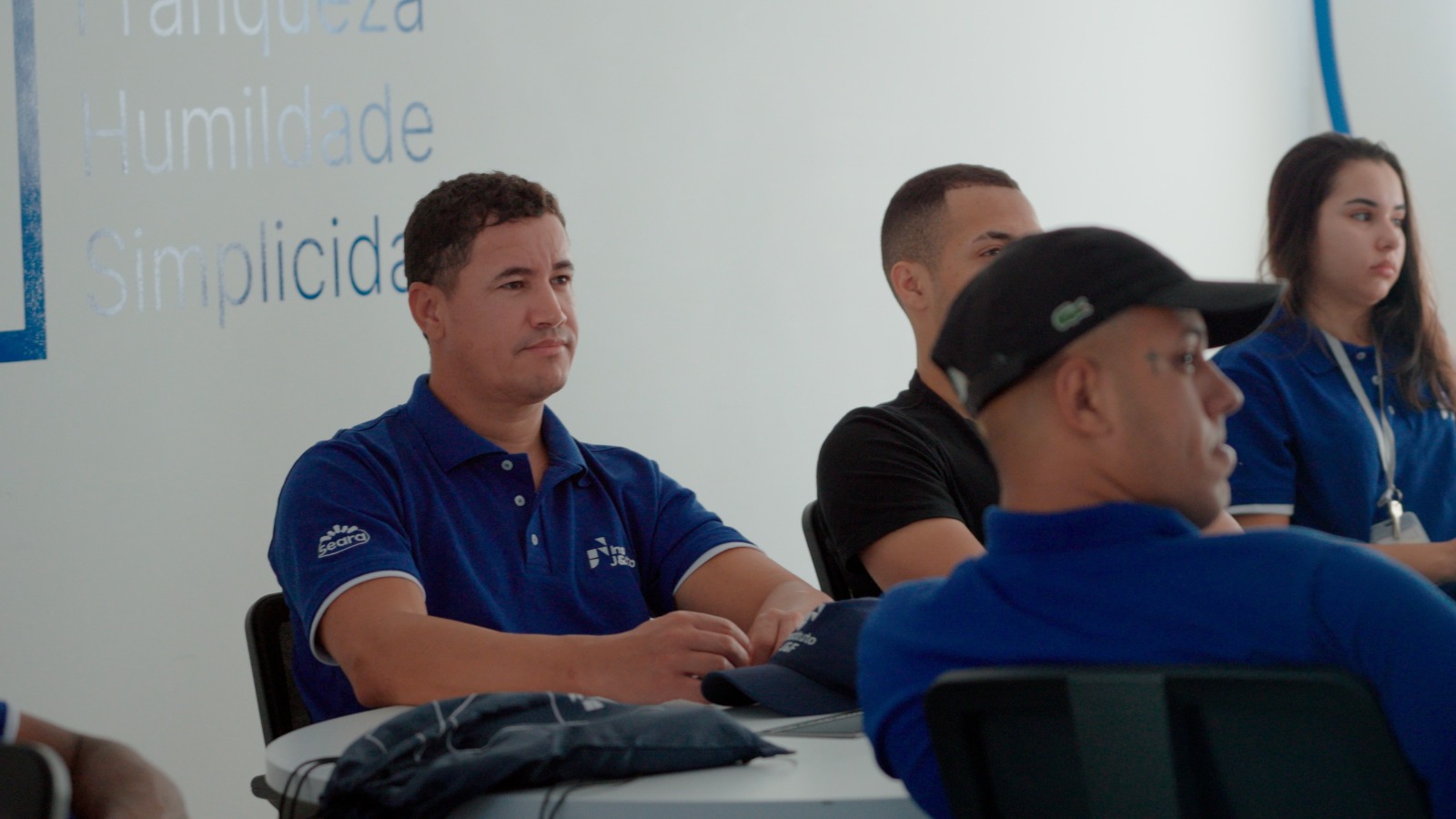 Alunos ouvindo em sala de treinamento com banner.