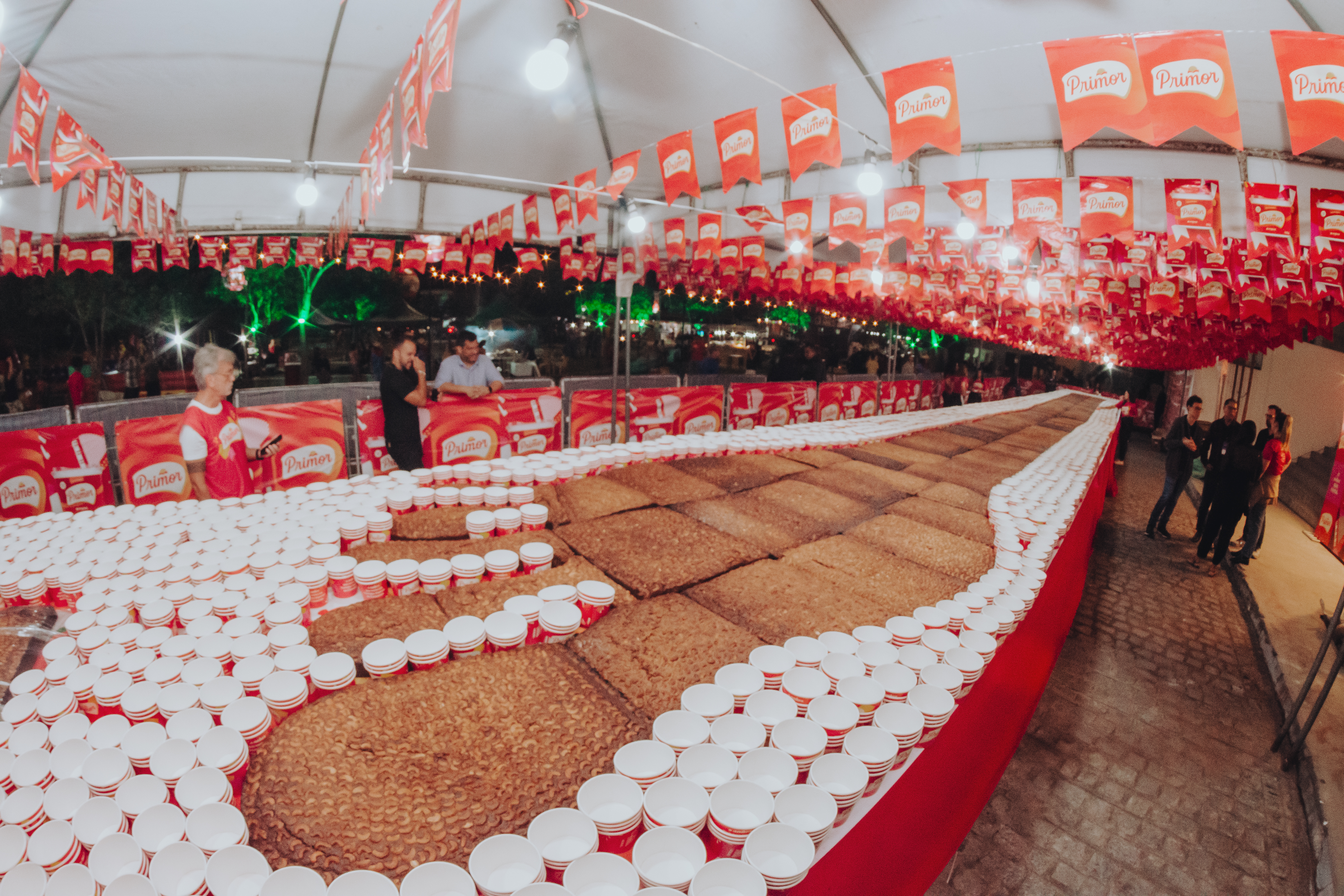 Imagem traz o doce pé-de-moleque em tamanho gigante sobre uma mesa enorme decorada com copos da marca Primor para degustação.