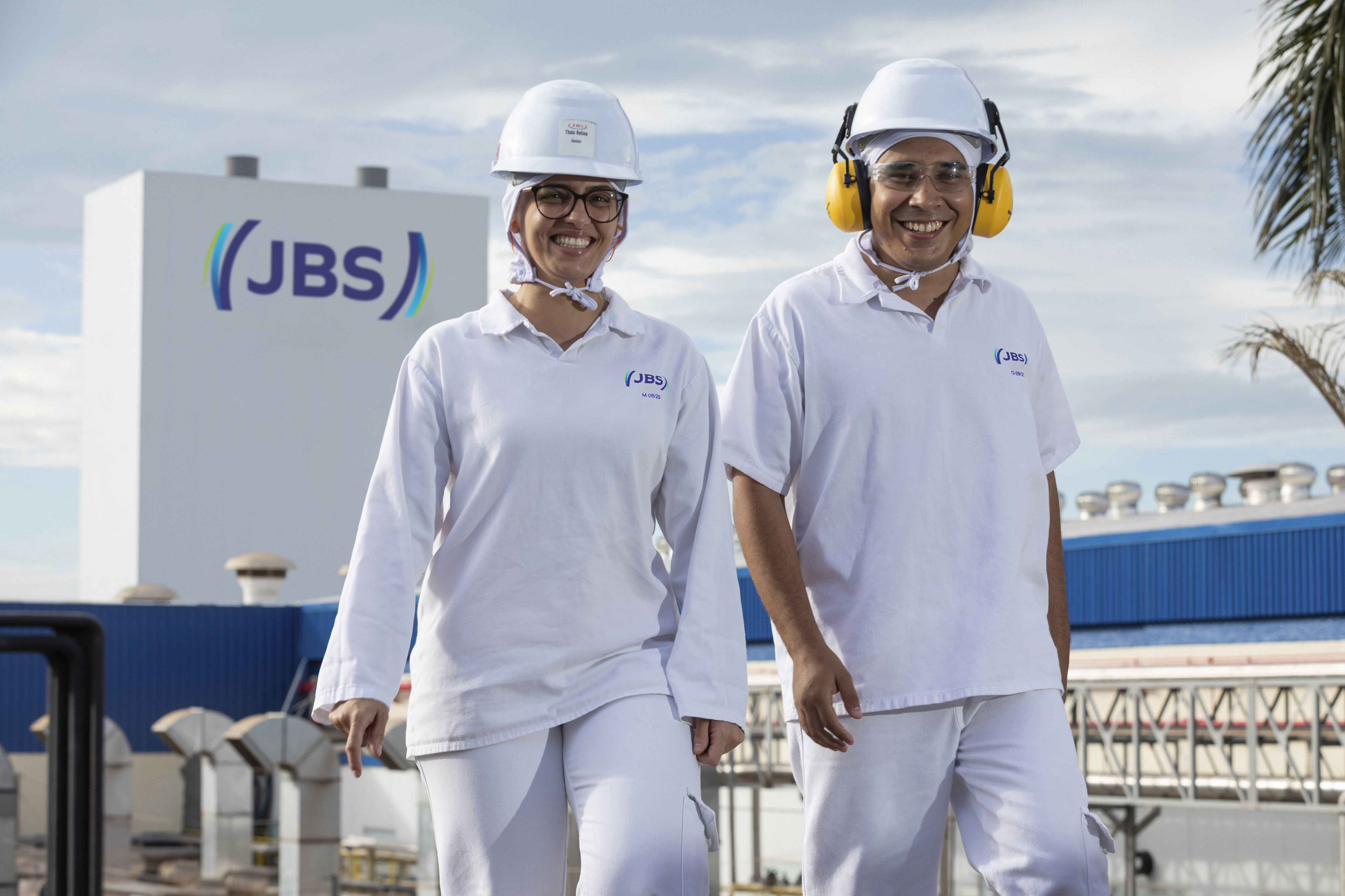 Em um cenário matutino, dois colaboradores JBS uniformizados, com capacete e óculos, sendo a mulher sem protetor auricular e o home com protetor. Ambos caminham em uma unidade JBS com uma torre com o logo da Companhia ao fundo