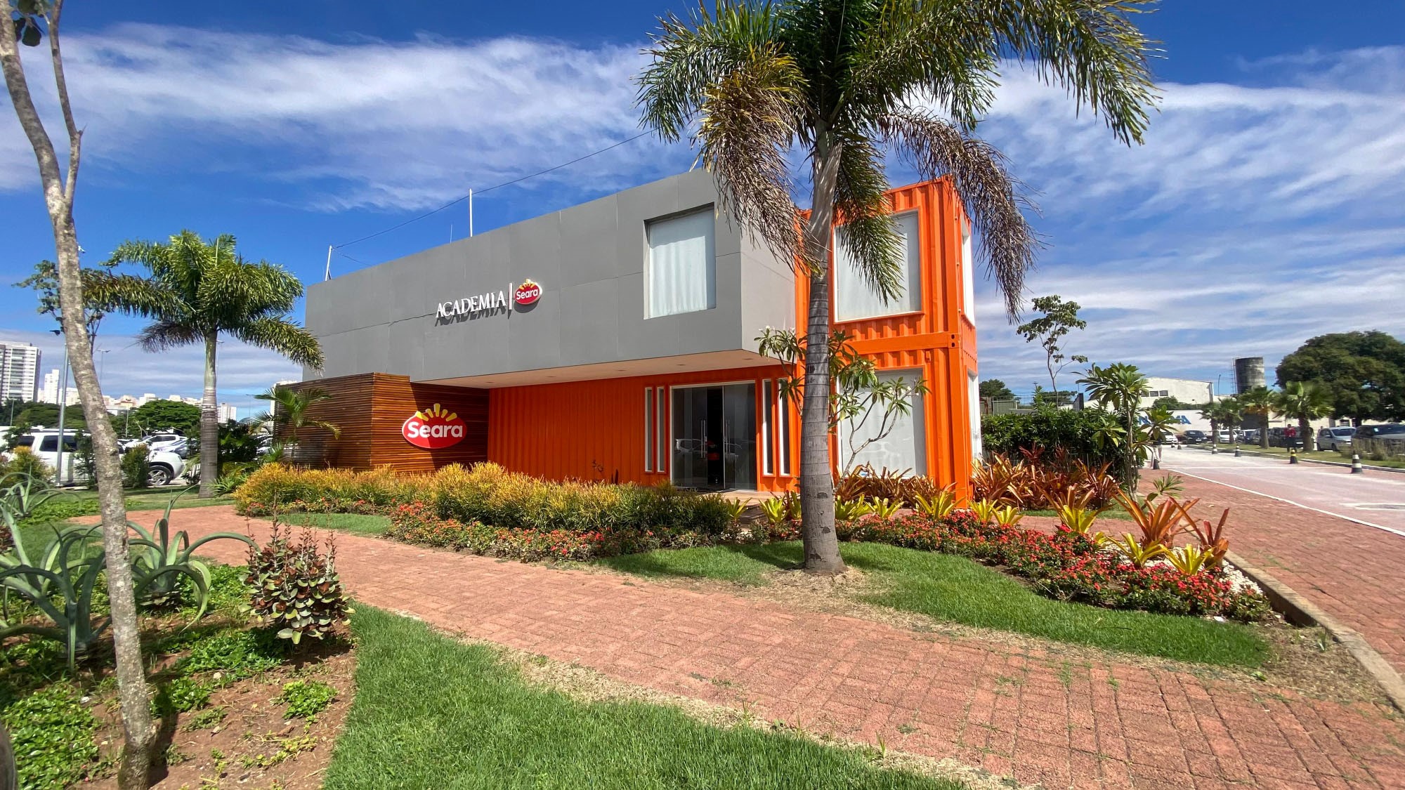 Uma construção moderna nas cores cinza, laranja e um pouco de madeira, de dois andares com marca Seara num estacionamento arborizado num dia ensolarado.