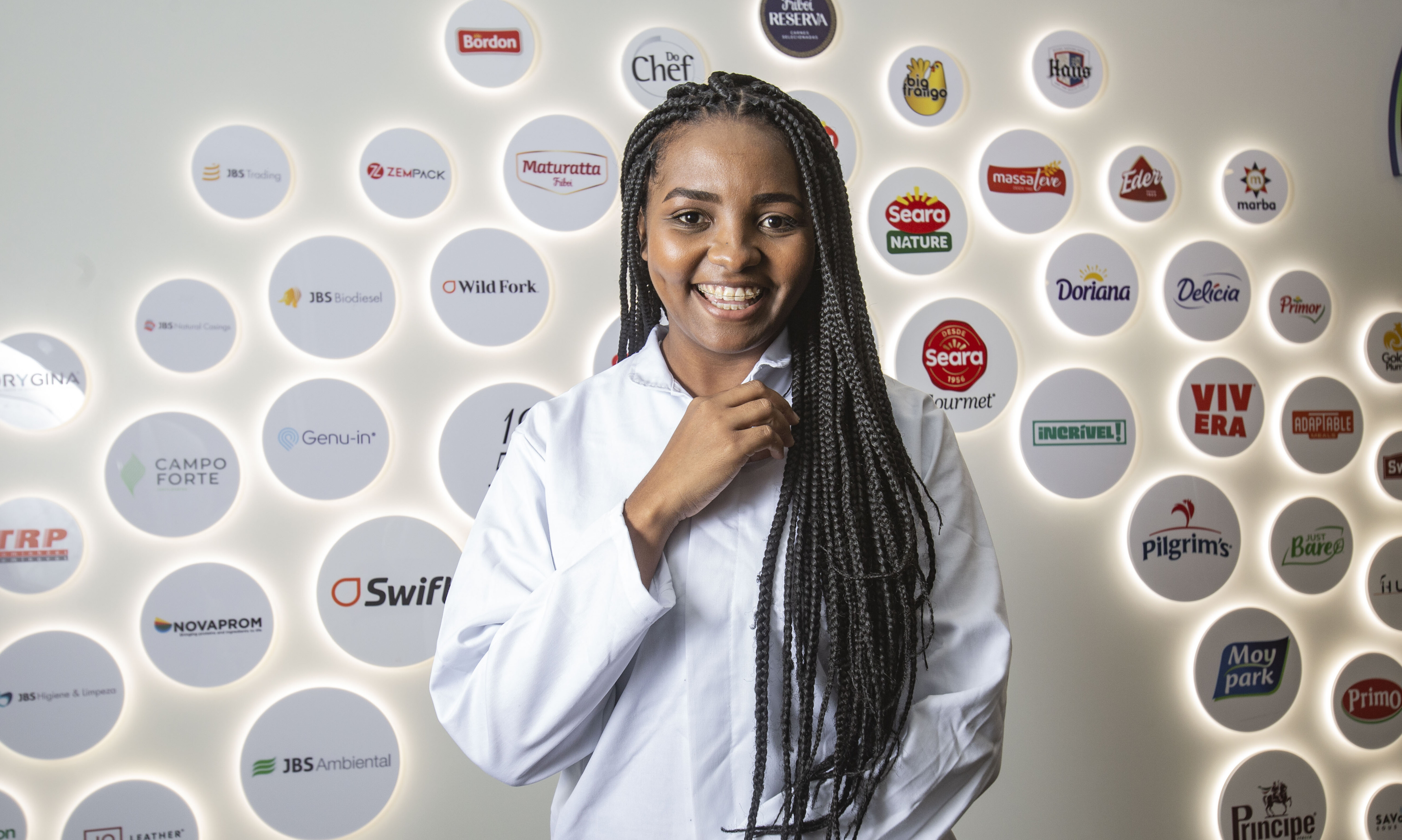 Num cenário com as logomarcas da JBS ao fundo, uma jovem colaboradora negra de pele preta com cabelos trançados, está uniformizada de branco segurando com sua mão esquerda a lapela do seu jaleco. A menina, que possui aparelho dentário, posa sorridente.