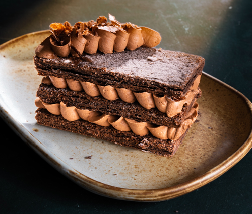 A imagem traz um prato de cerâmica em formato oval na cor azul envelhecida com manchinhas marrons, sobre o prato, uma fatia de torta folhada de chocolate em três camadas com recheio de ganache de chocolate em formato de ondas fazendo um volume artístico. Por cima, um detalhe como o recheio dando o toque final no topo da fatia.