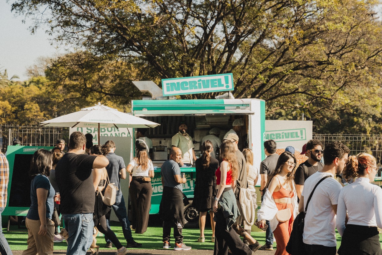 Em um dia ensolarado num cenário de parque com um gramado um food truck branco e verde com um letreiro escrito INCRÍVEL e pessoas transitam em frente, algumas aguardam na fila e um deles é atendente que está no balcão.