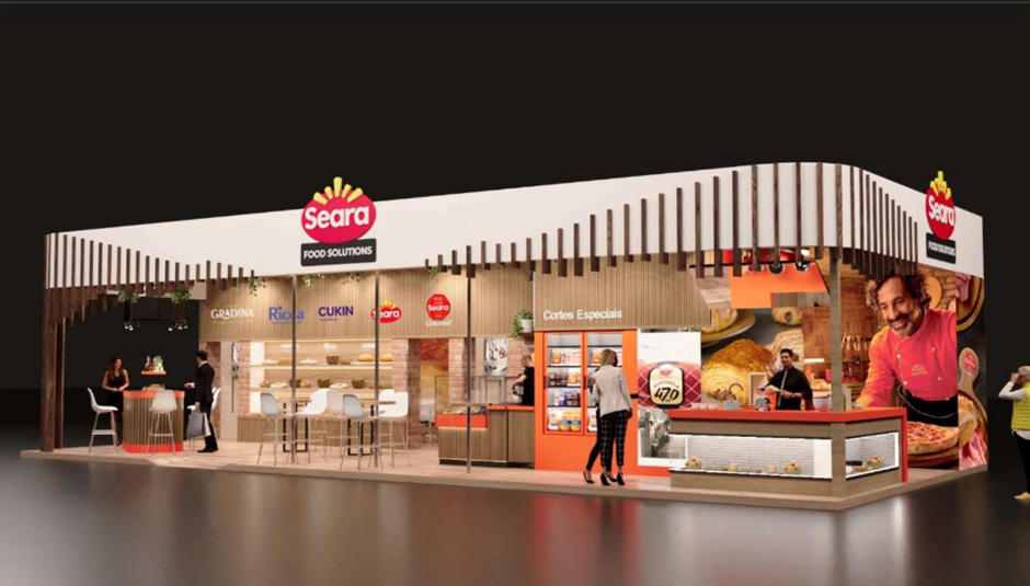 Imagem traz a projeção do stand na feira com a marca Seara Food Solutions destacada numa estrutura de madeira clara que traz um balcão de padaria geladeiras e imagens de produto como mortadela, pães e um cozinheiro com uma pizza que acaba de sair do forno em mãos. No stand também tem mesas com cadeiras e as marcas do grupo que participam da exposição, como Gradina, Ricca, Cukin Seara e Seara Gourmet.