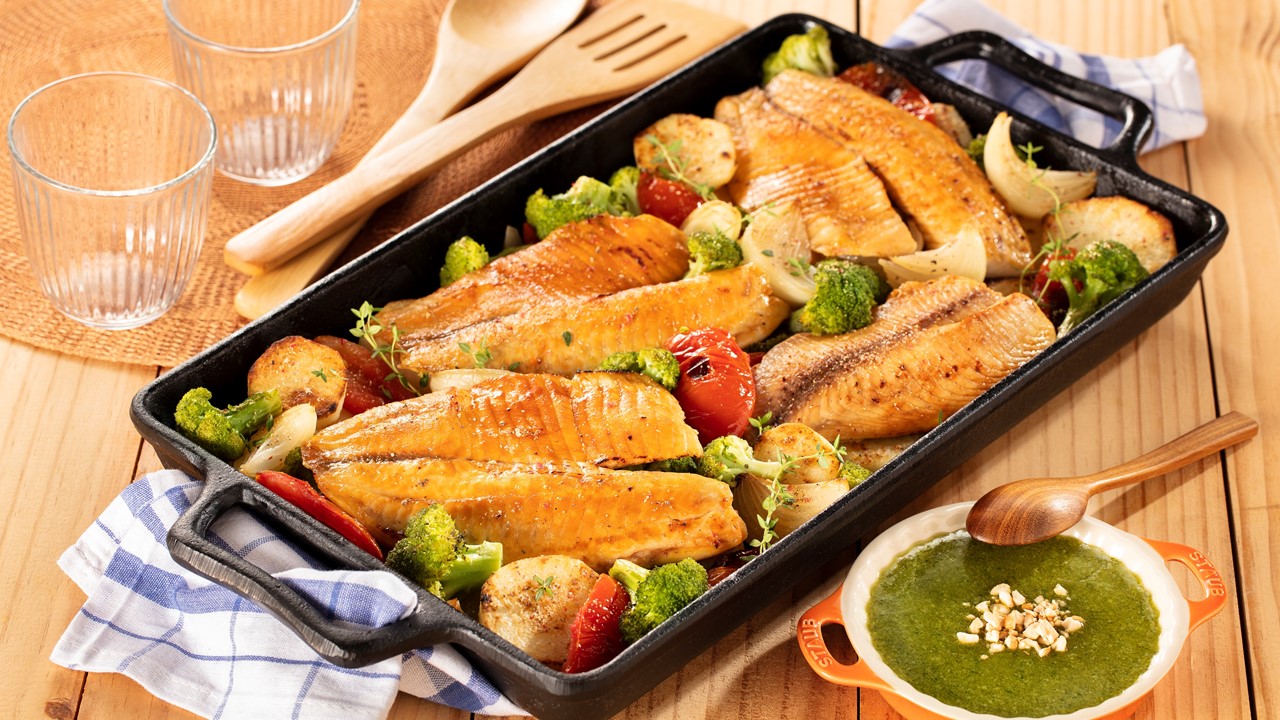Bandeja de filé de Tilápia sobre mesa de madeira, ao lado de recipiente com molho pesto, copos de vidro e garfo e colher de servir.