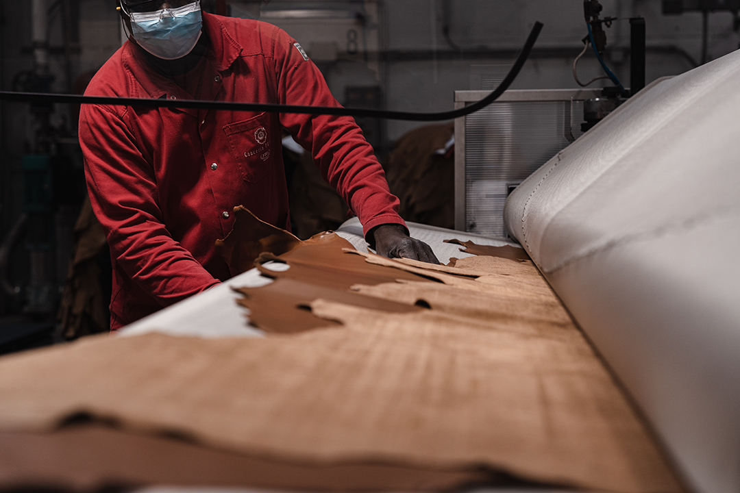 Trabalhador está com uma roupa vermelha, posicionado próximo a uma máquina com uma peça de couro sendo manipulada